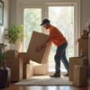 Man in orange shirt carrying a large cardboard box indoors. Boxes are stacked near the window.