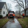 Two men loading brush into a red dump truck parked on a driveway between two houses.