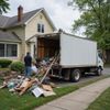 Man loading debris into a moving truck parked in front of a yellow house.