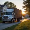 A white garbage truck parked on a suburban street with sunlight. A person is near a car.
