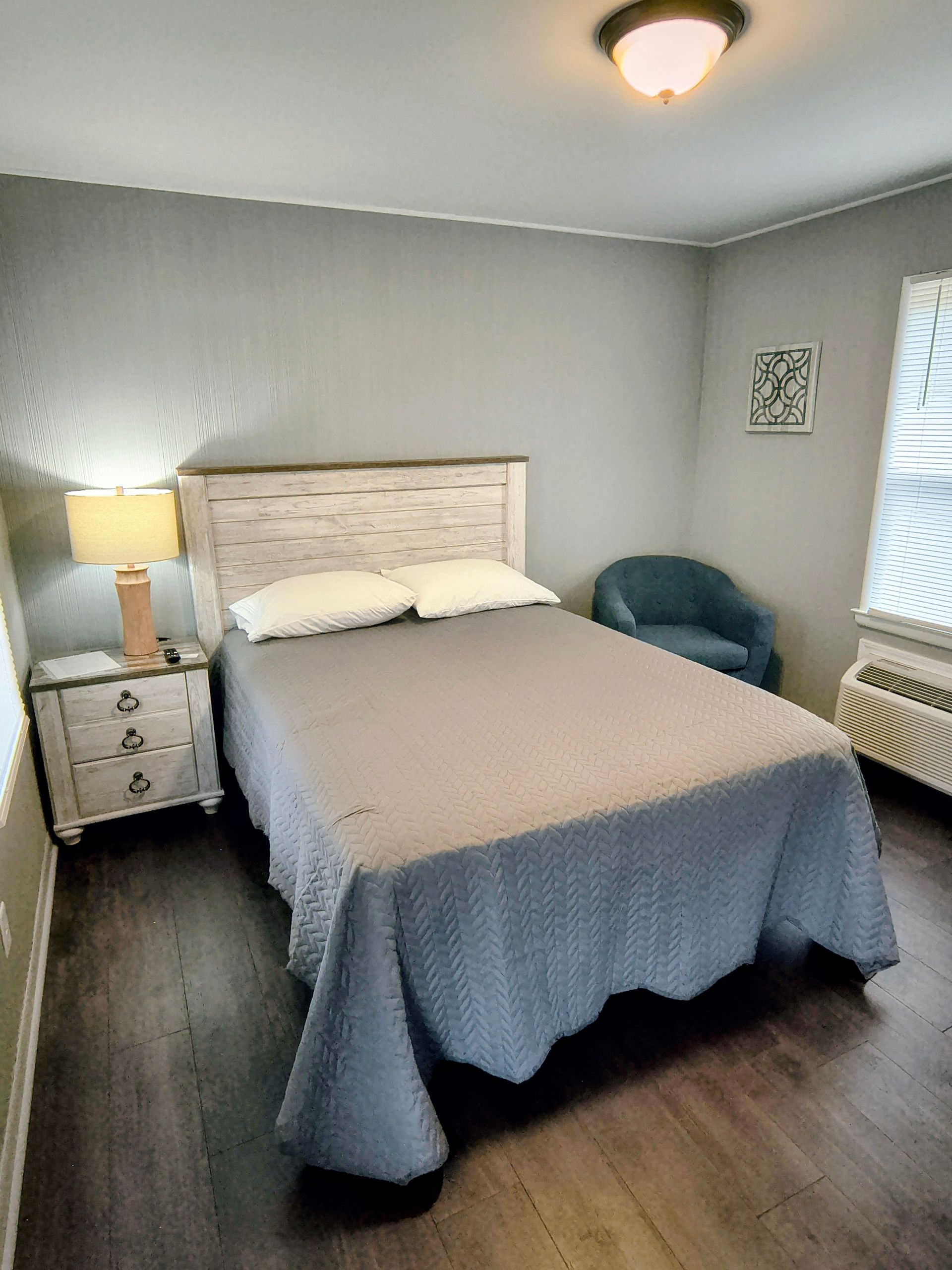 Bedroom with queen bed, nightstand with lamp, small armchair, and window with blinds.