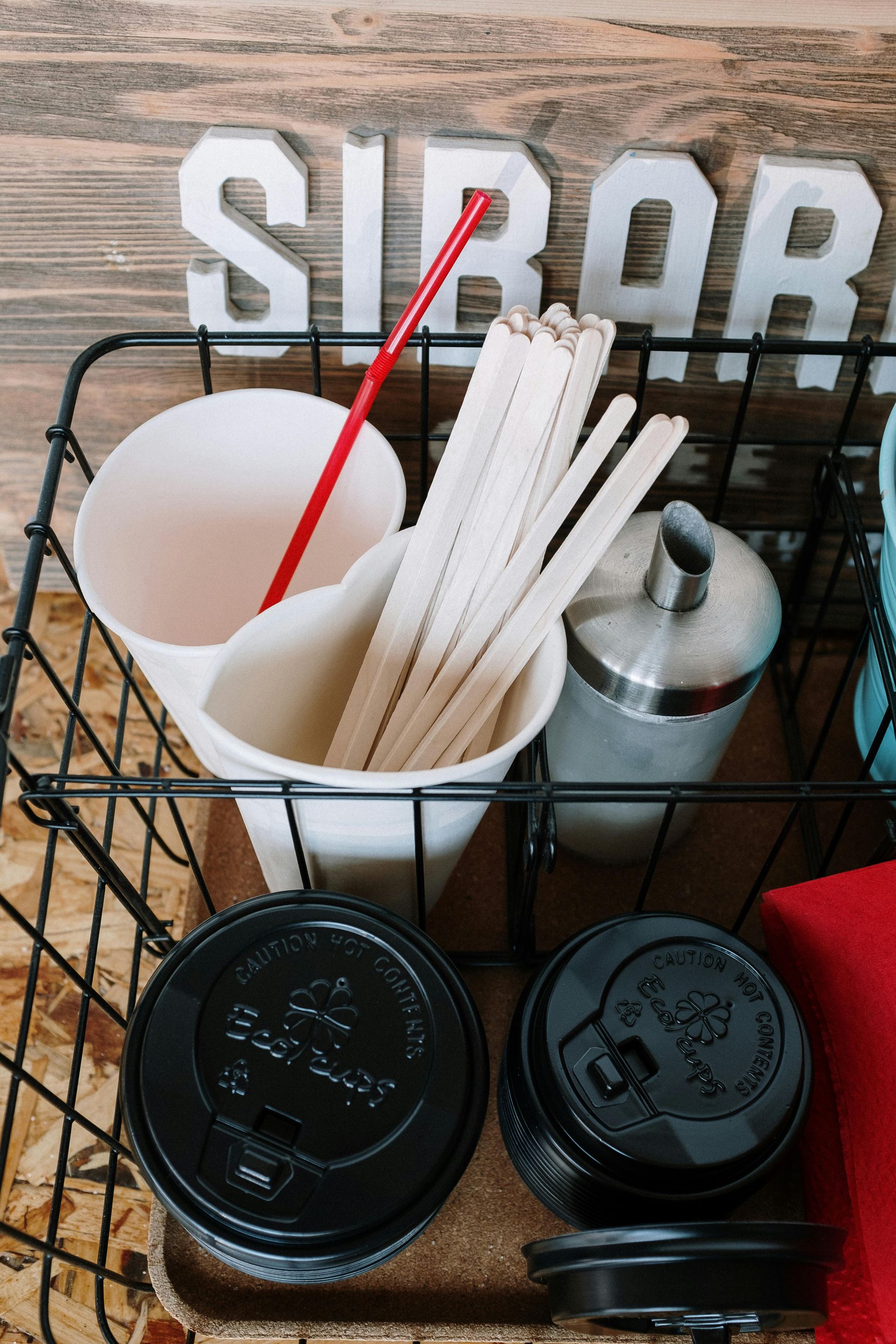 A black wire basket contains white cups, lids, stir sticks, a red straw, and a metal sugar dispenser .