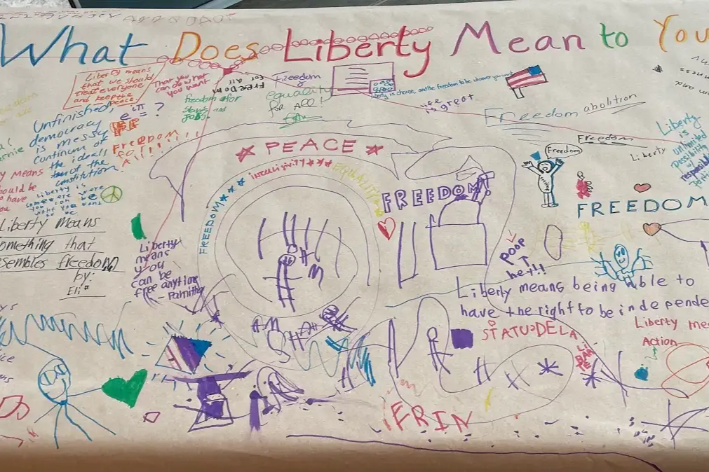 Woman assisting a child at a table outdoors; other people in the background.