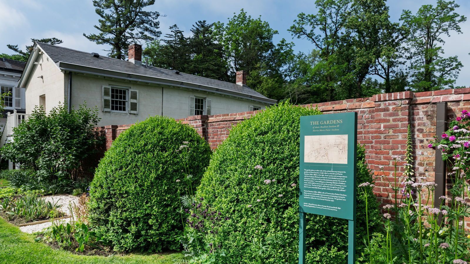 A white building behind a green garden, a brick wall, and a sign.