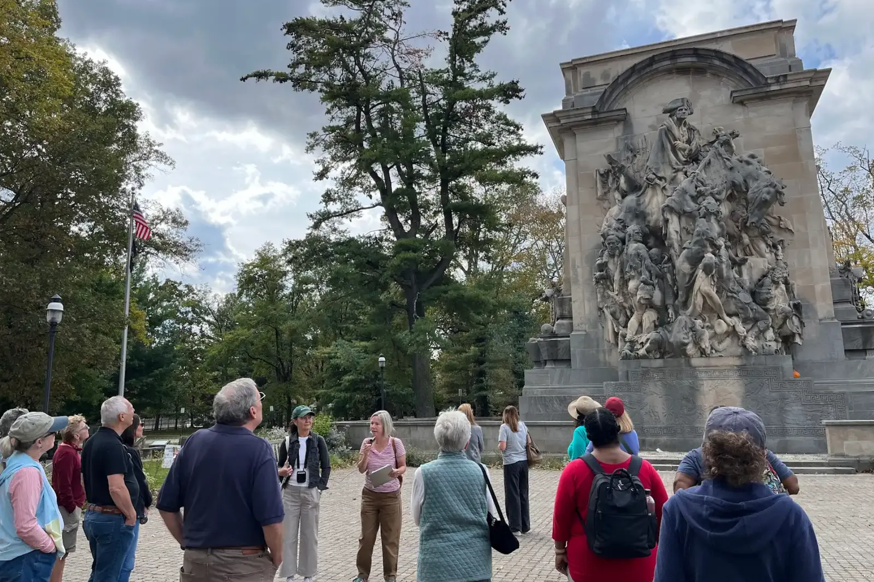 Group of people looking at a large sculpture, possibly on a tour. Cloudy day, trees in background.