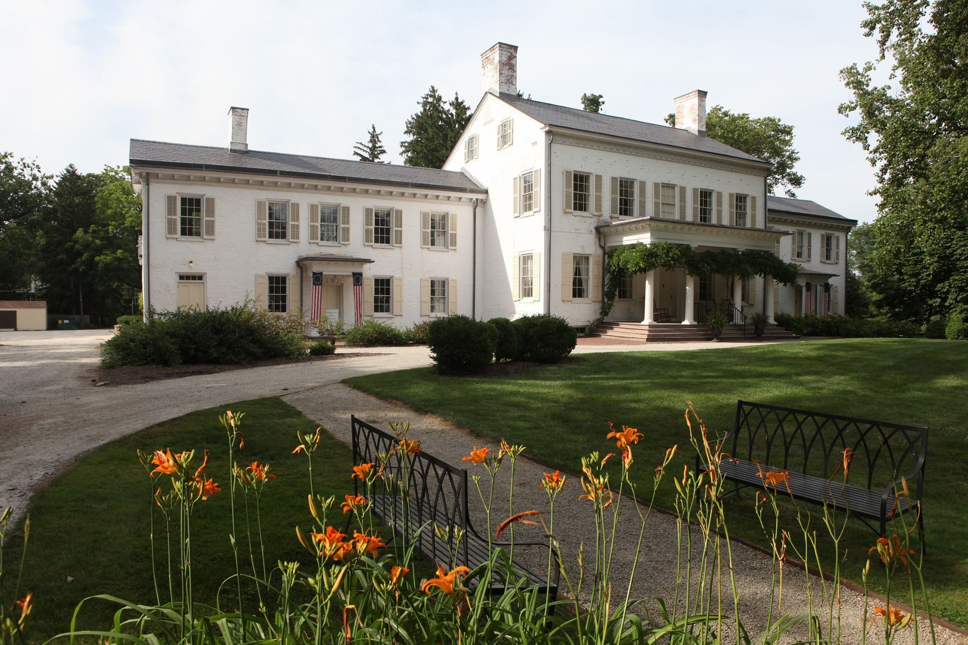 White mansion with columns and shutters, path leading through garden with orange lilies, bench.