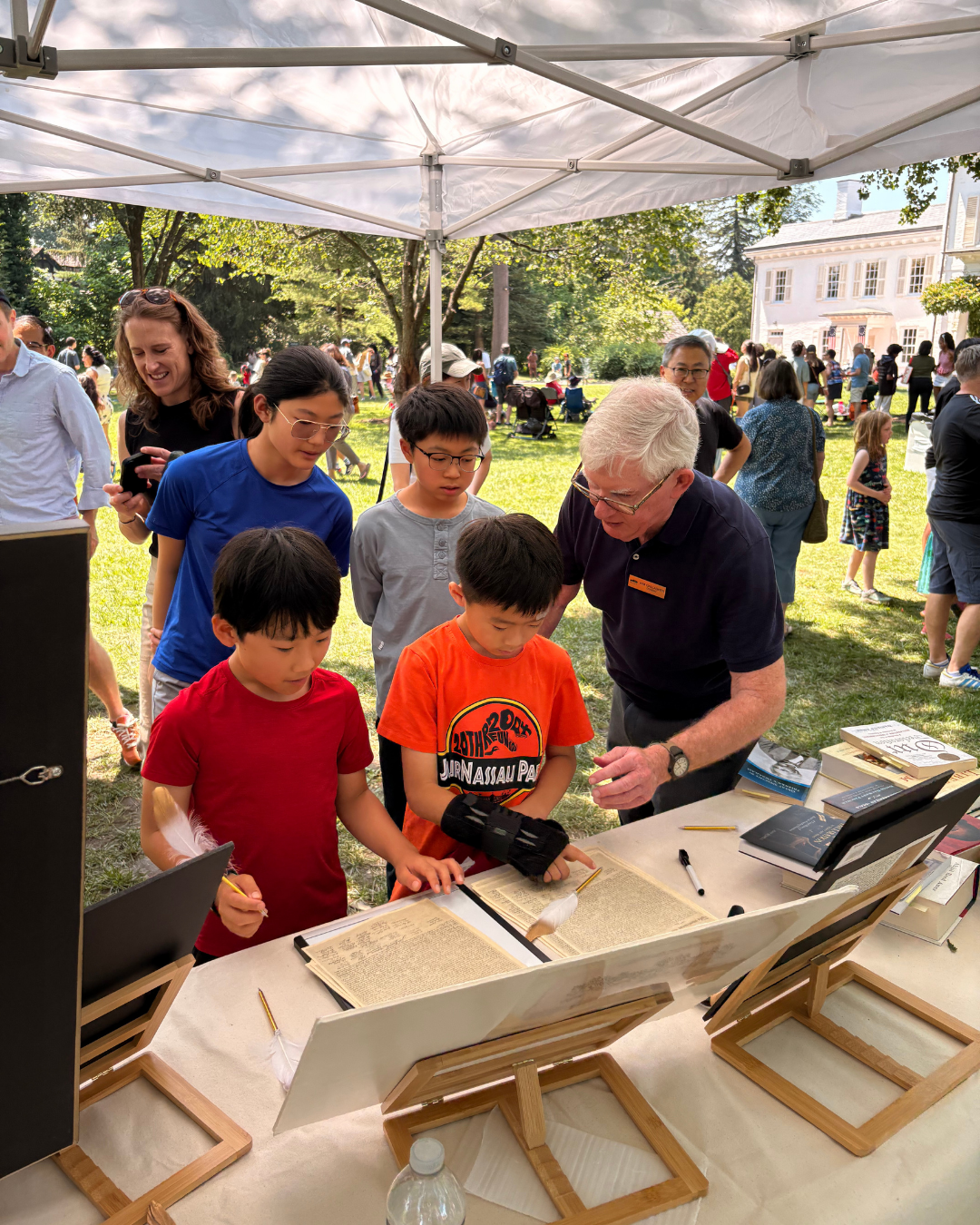 Children and adult looking at a display on a table outside. People gather on grass with a white tent.