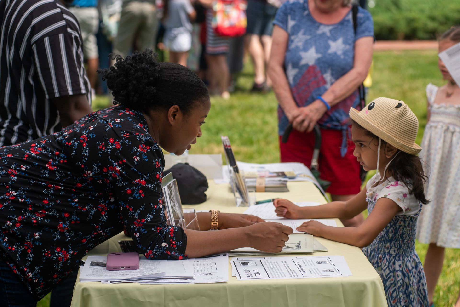 Woman assisting a child at a table outdoors; other people in the background.