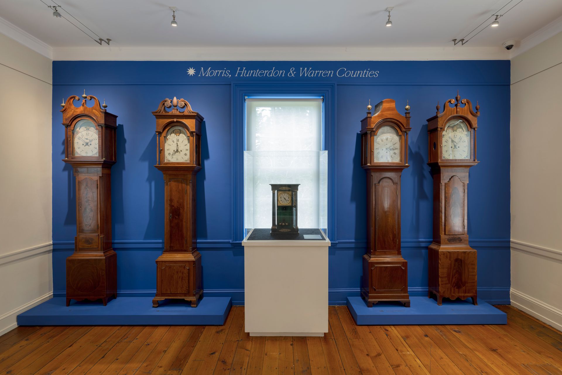 Five tall clocks in a room with blue walls. A smaller clock is in the center. Wooden floor.