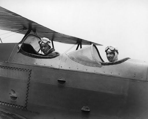 Two pilots in an open cockpit biplane, wearing goggles and looking forward.