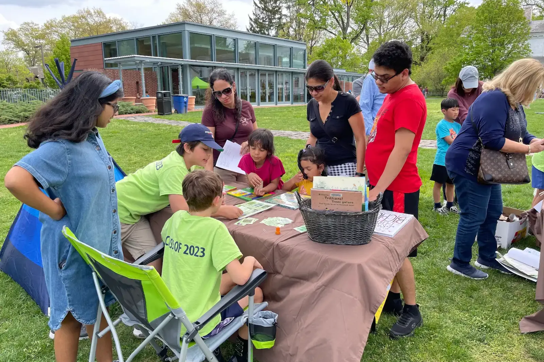 People at an outdoor table; children and adults playing a game; green lawn; glass building in background.
