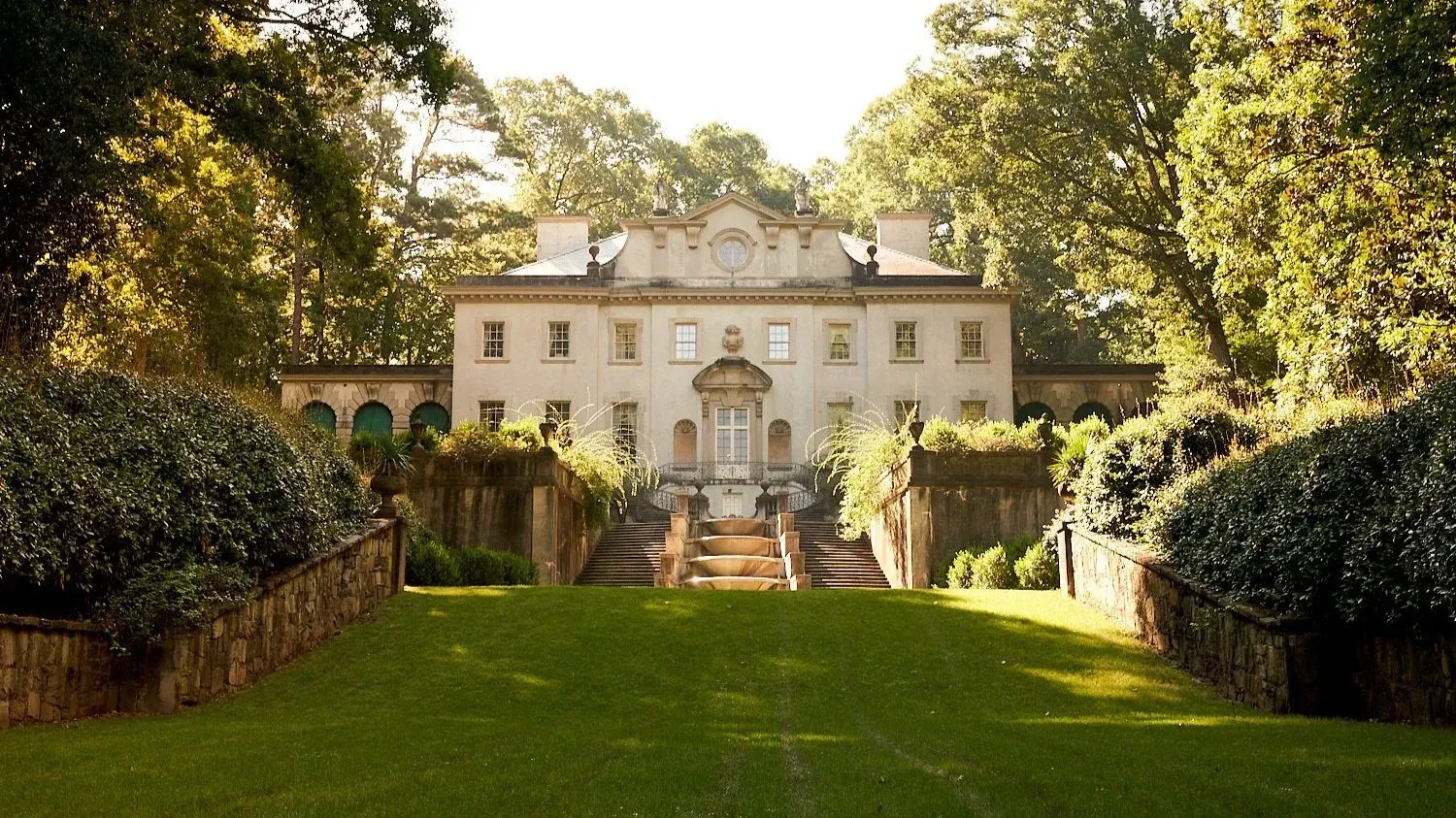 A large, light-colored mansion surrounded by greenery with a grand staircase and lawn.
