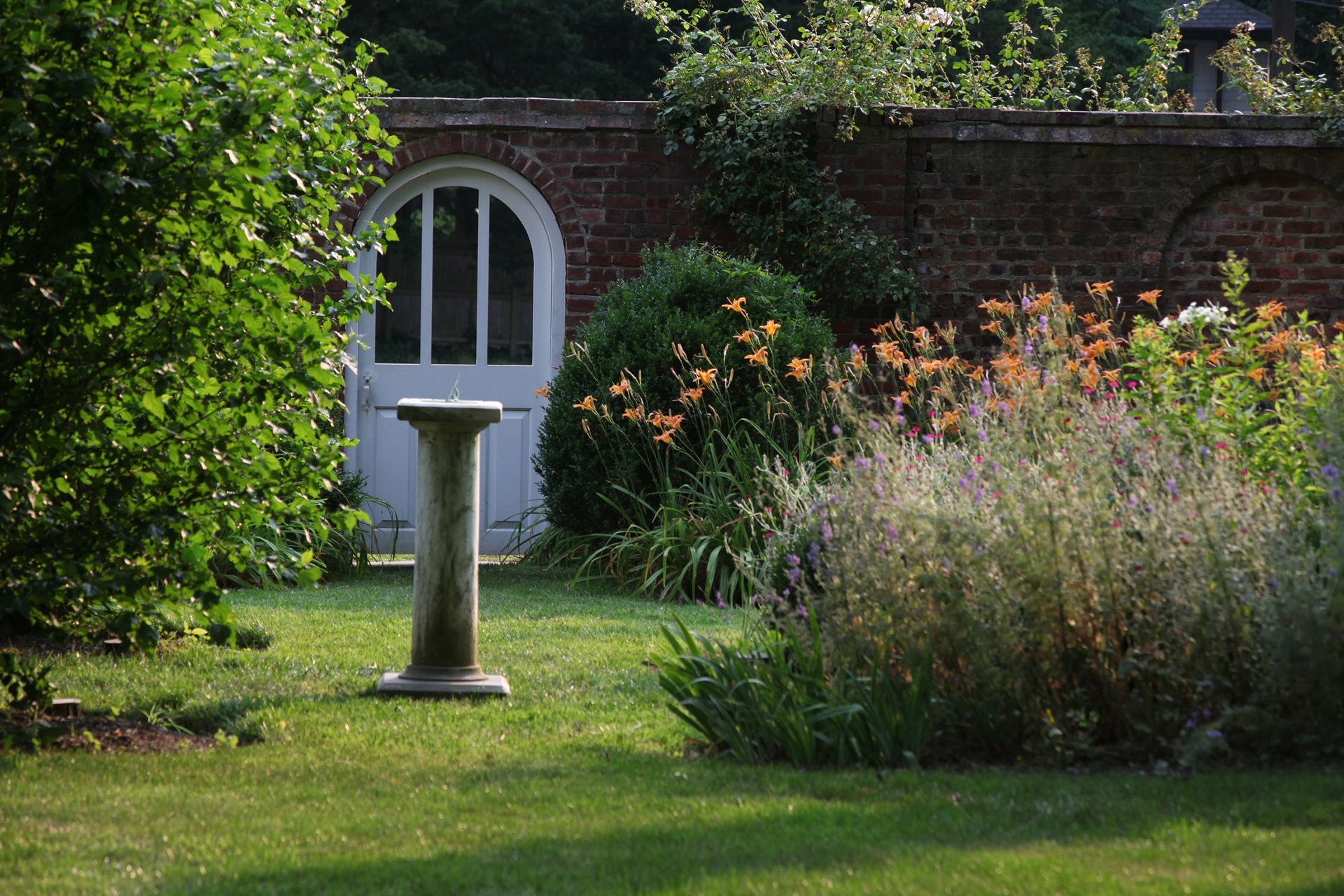 Lush garden with white arched door, stone pedestal, and flowering bushes against a brick wall.