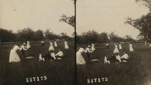 People in white dresses and suits in a grassy field. Some sit, others dance, under trees.