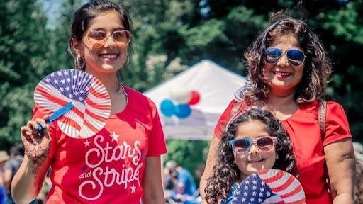 Three women smiling at an outdoor event, holding American flags and fans, wearing red, white, and blue.