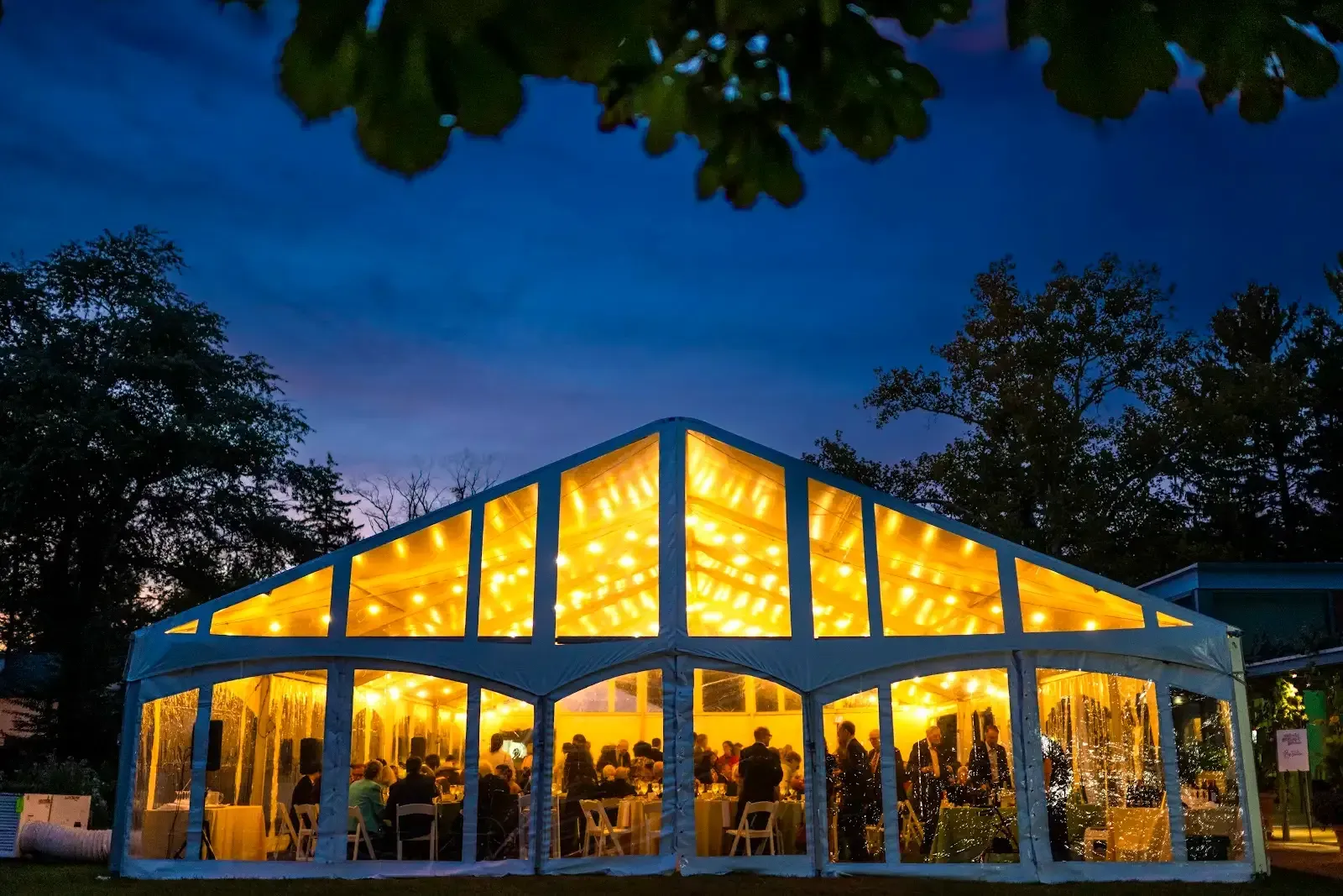 Illuminated tent at dusk hosting a social gathering with people dining and socializing.