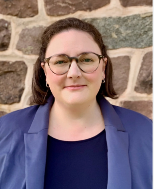 Portrait of a person wearing glasses and a blue blazer, standing against a stone wall.