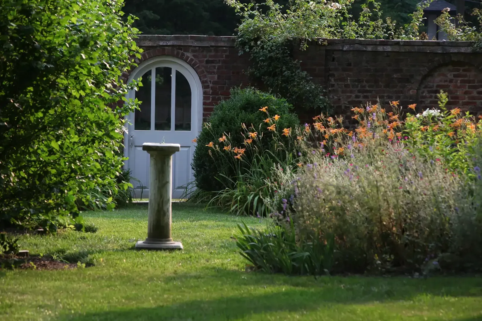 Garden scene: White arched door in brick wall, pedestal, green plants, orange flowers, sunny.