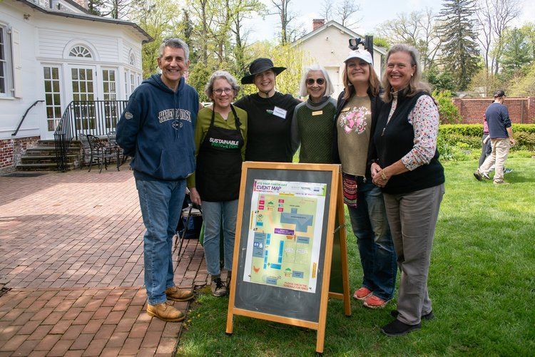 Group of people posing by a map in a garden; sunny day, smiles, trees, and a white house in background.
