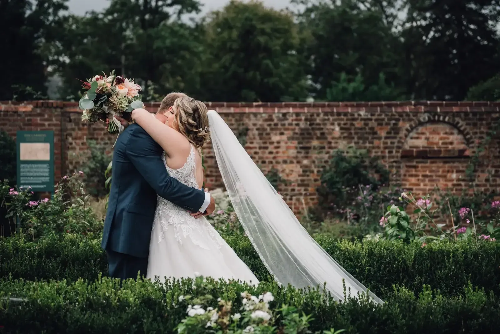 Bride and groom embrace in a garden. The groom wears a blue suit, the bride a white dress and veil. Red brick wall in background.