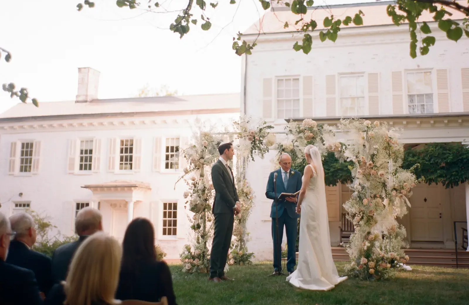 Wedding ceremony outdoors with couple, officiant, floral arch, and white building.