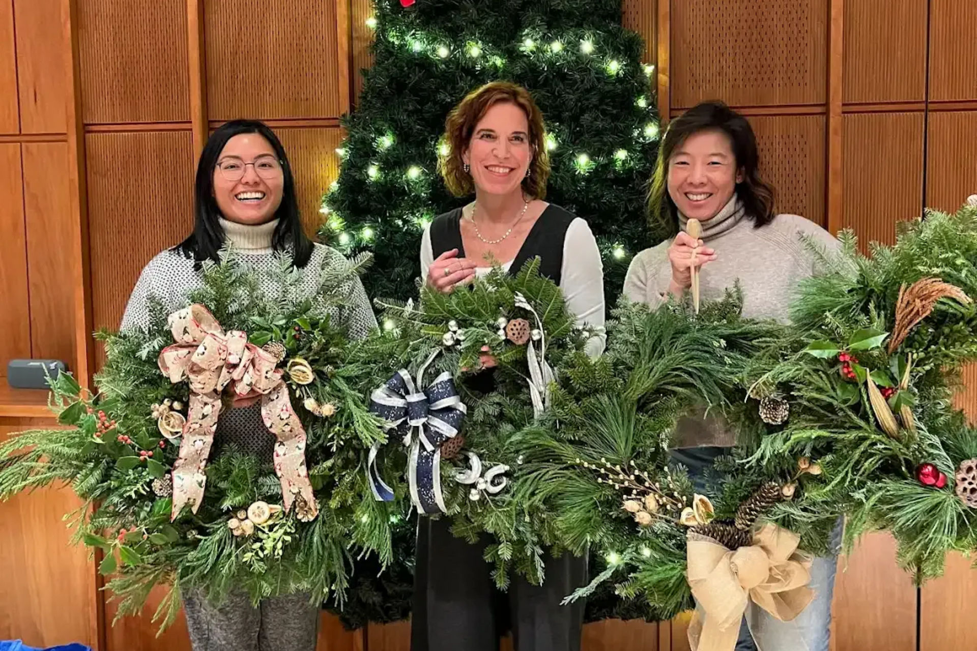 Three people holding decorated wreaths in front of a Christmas tree.