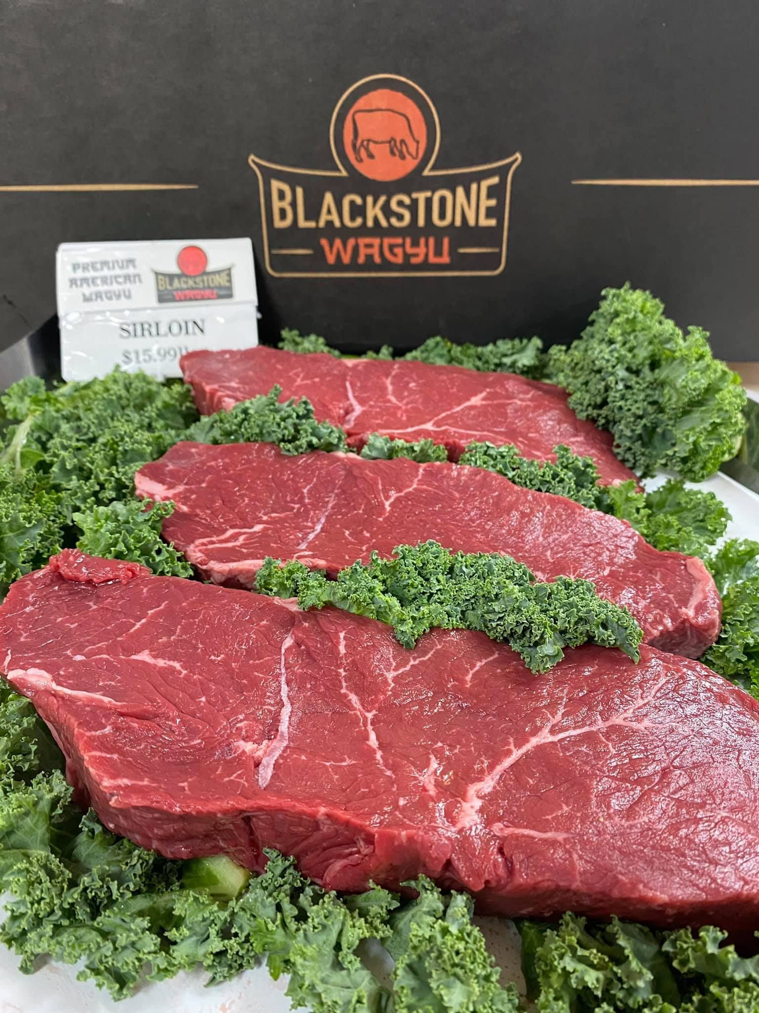 Three raw Blackstone Wagyu sirloin steaks on a bed of kale, with the brand logo in the background.