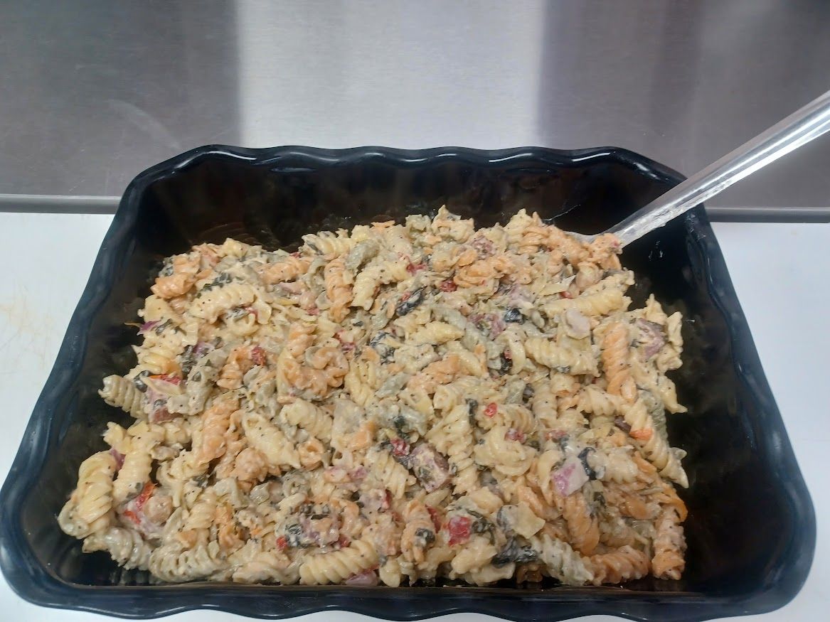 Pasta salad in a black tray, with a silver spoon. The salad has visible herbs and red vegetables.