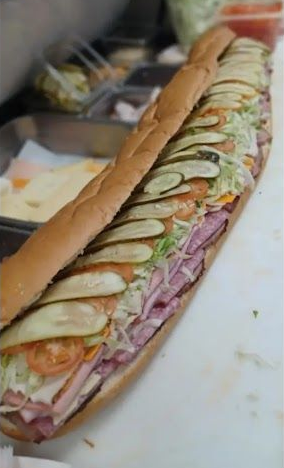 Long submarine sandwich with pickles, tomatoes, lettuce, and deli meats on a white surface.