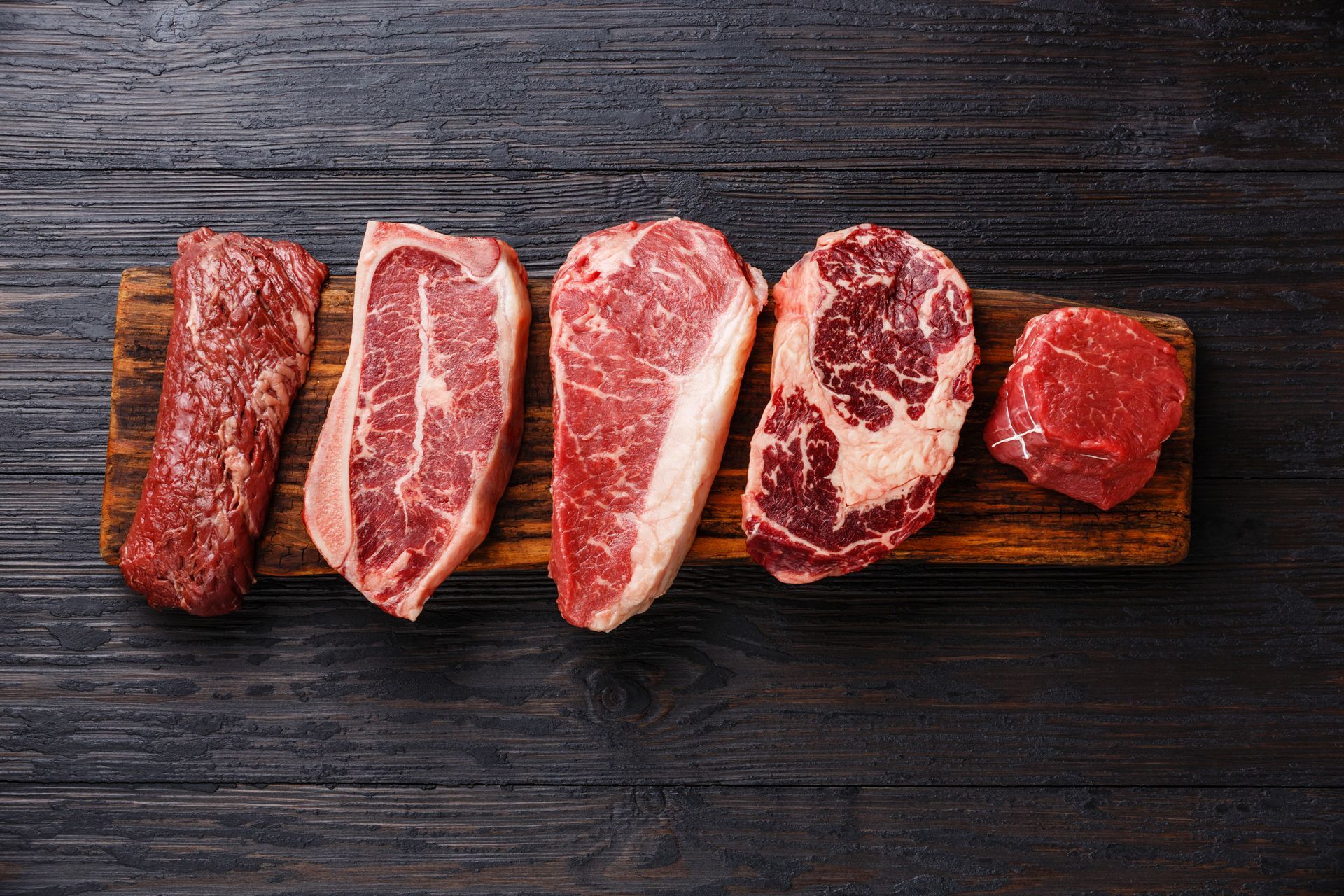 Five raw steak cuts on a wooden board atop a dark wooden surface.