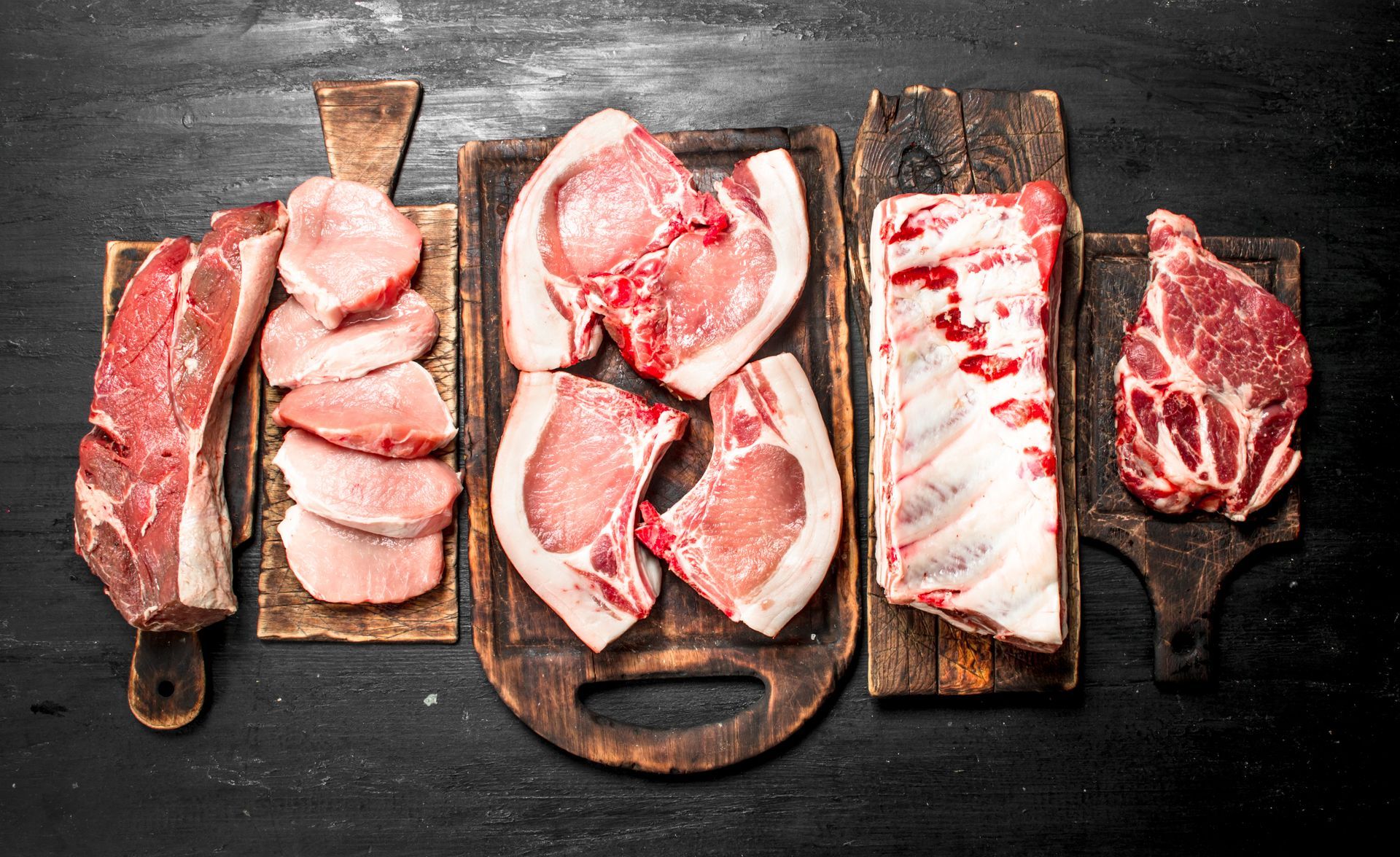 Various cuts of raw meat arranged on wooden cutting boards against a black background.