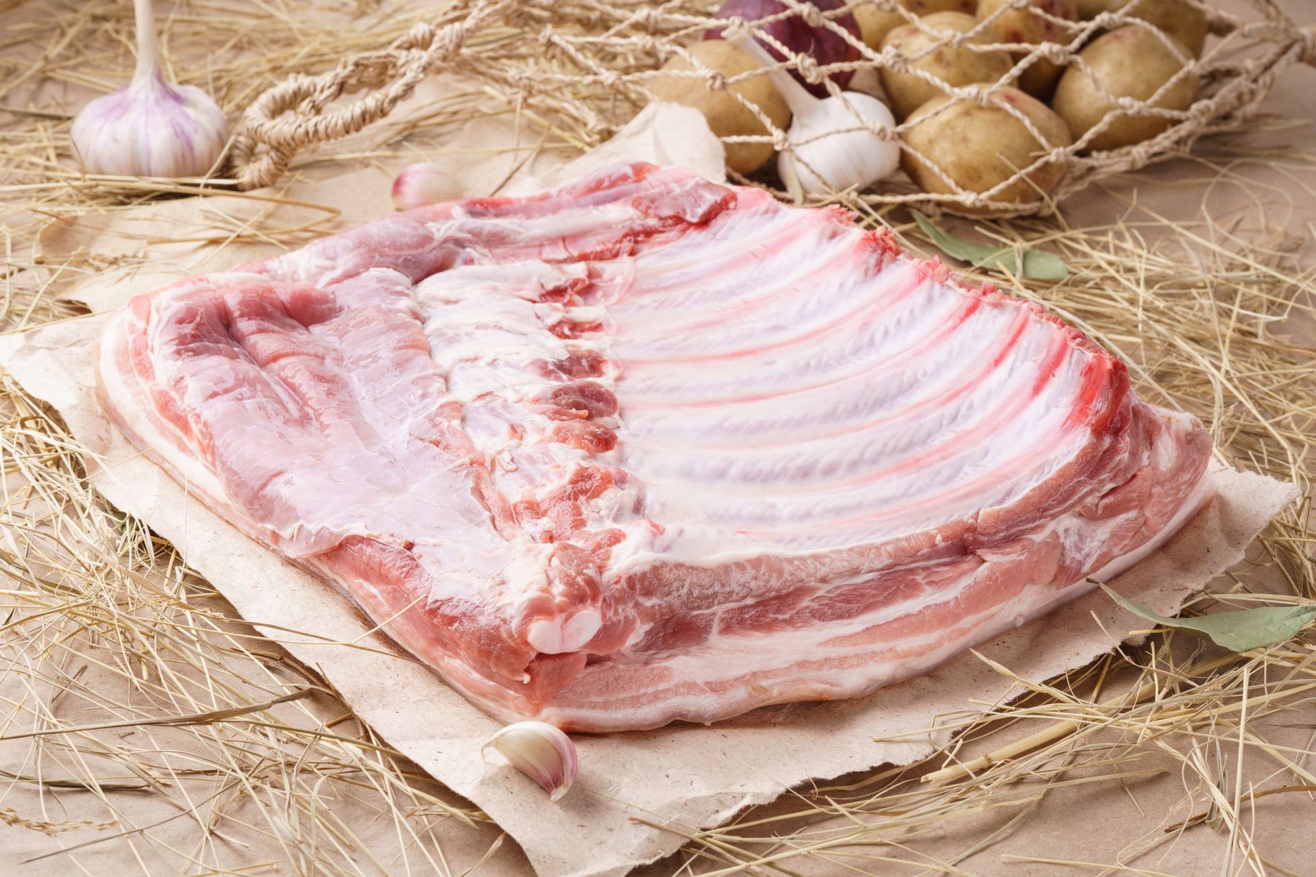 Raw pork belly on butcher paper, with garlic and potatoes in background.