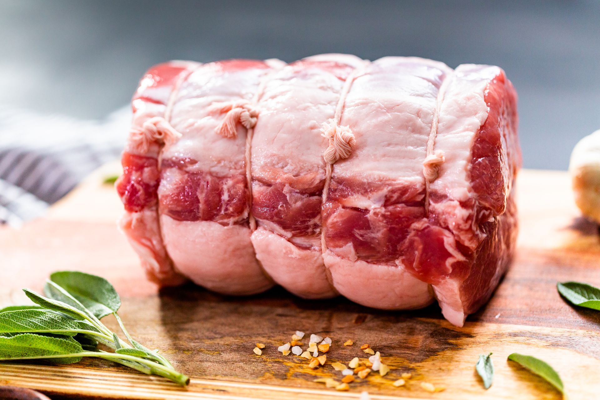 Raw pork roast tied with twine on a wooden board with herbs and seasonings.