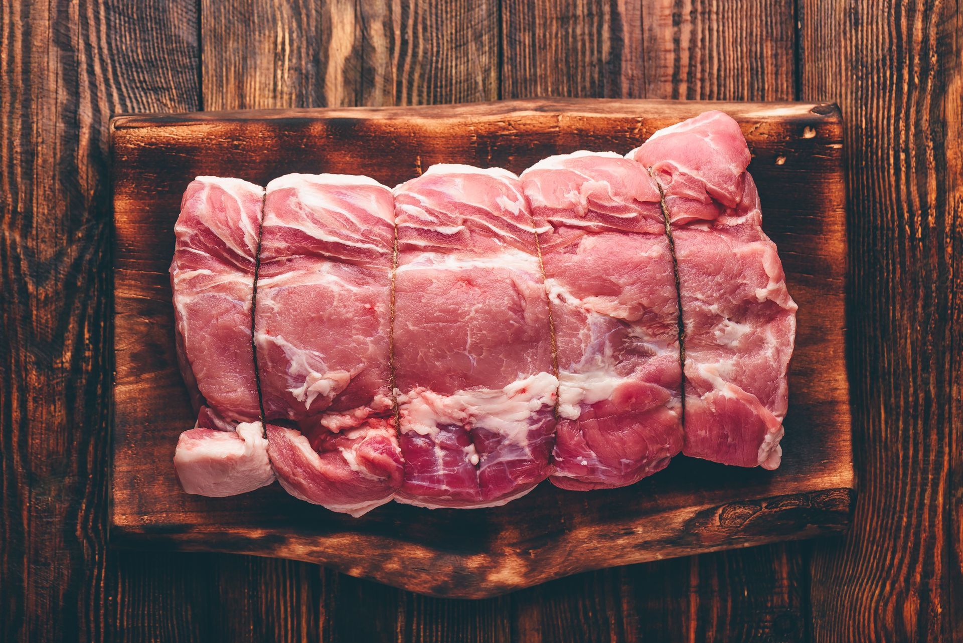 Raw pork roast tied with twine on a wooden cutting board, resting on a wooden table.