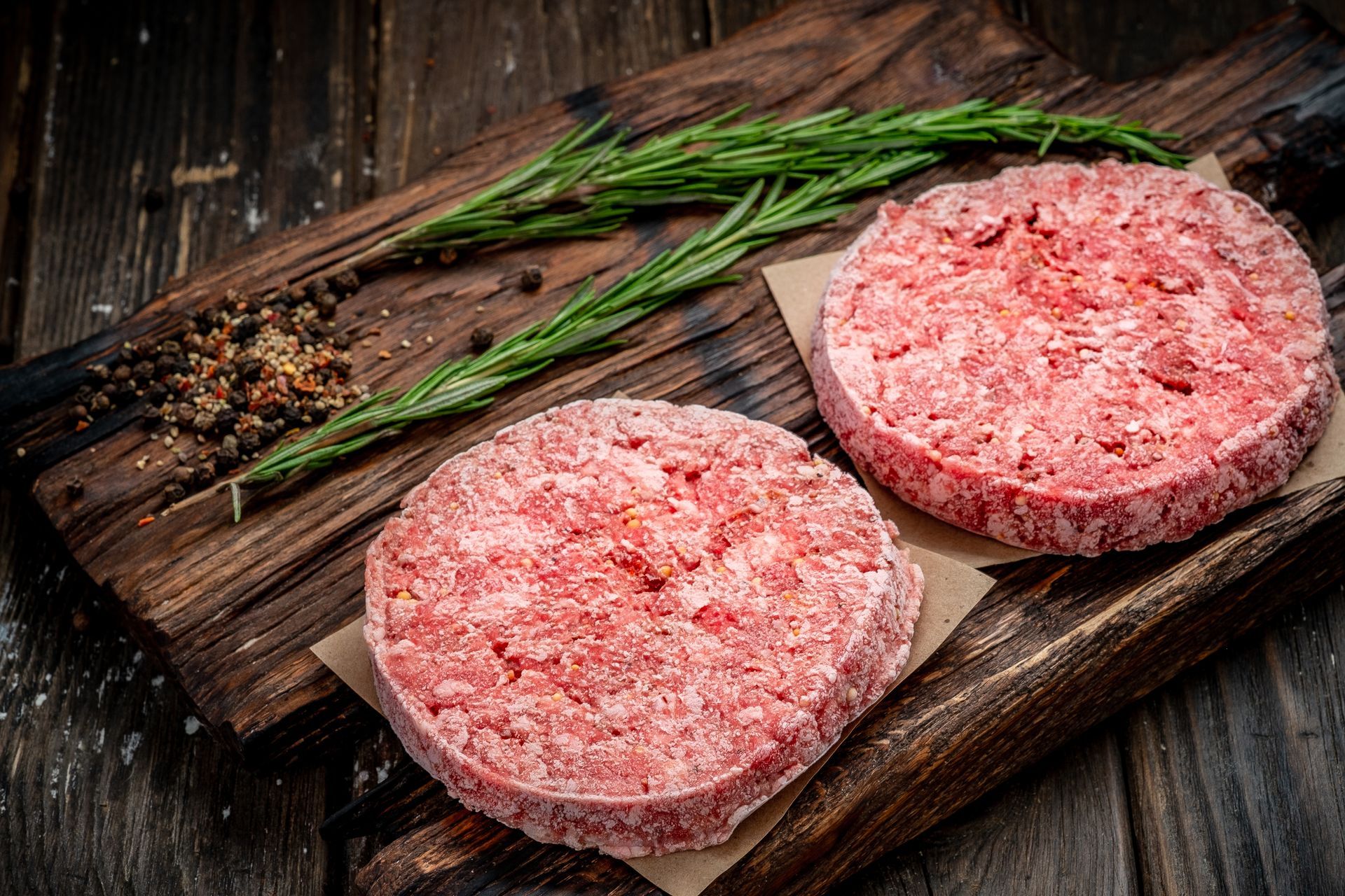 Two raw hamburger patties on a wooden board with rosemary sprigs and peppercorns.