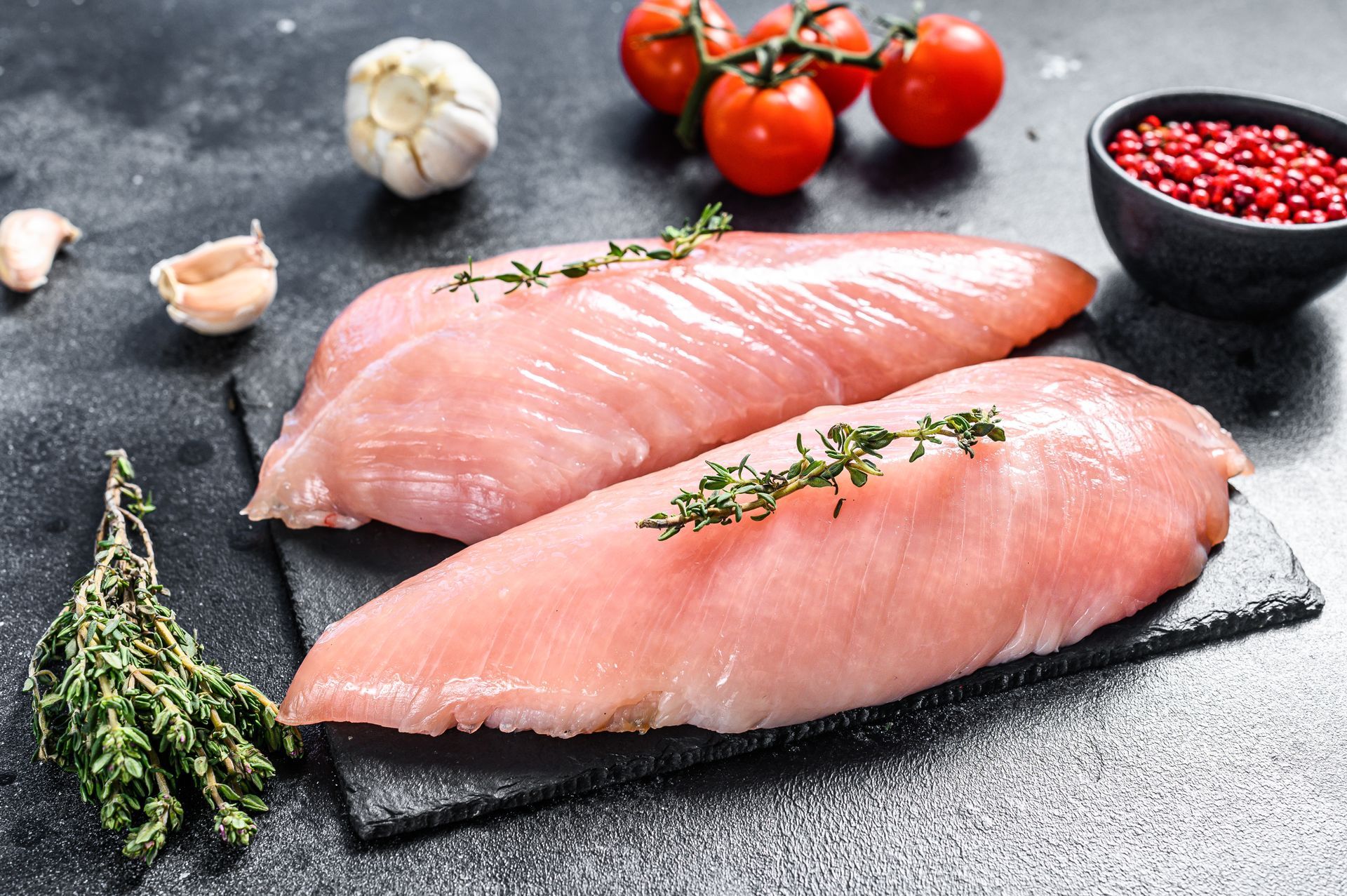 Two raw turkey breasts on a black slate, with tomatoes, garlic, thyme, and peppercorns.