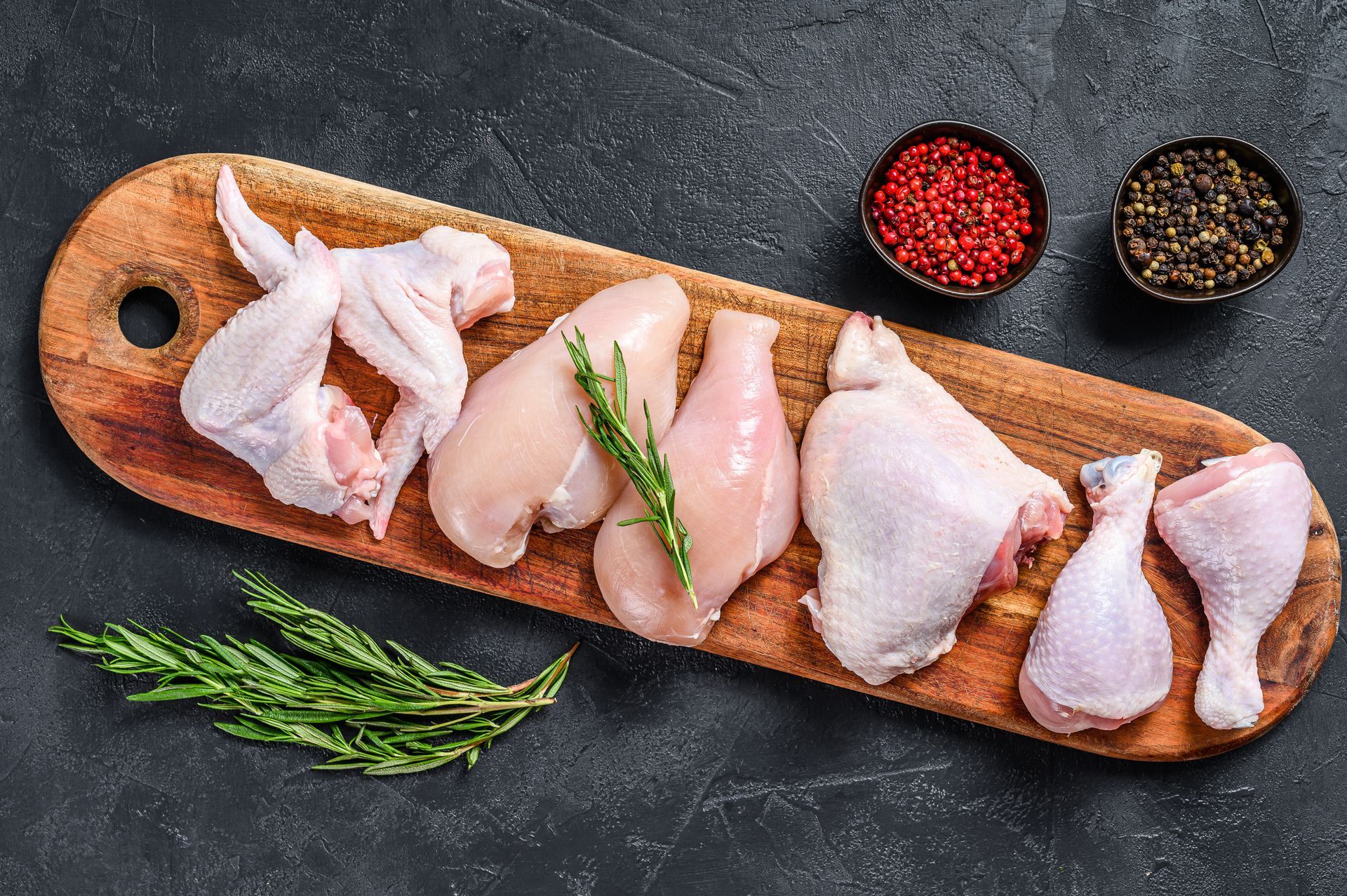 Raw chicken cuts arranged on a wooden board with herbs and spices on a dark surface.