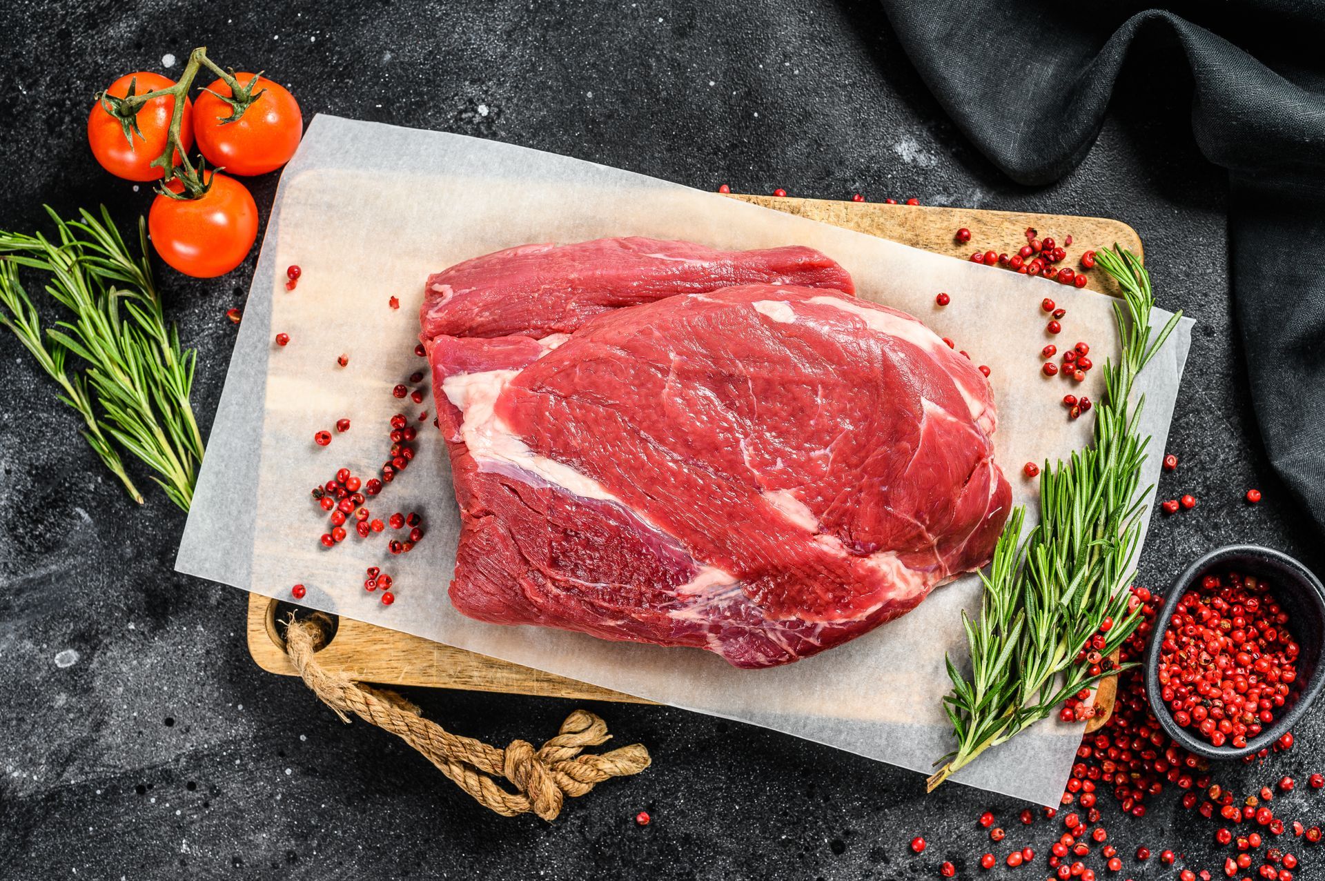 Raw beef roast on parchment paper, surrounded by tomatoes, rosemary, and peppercorns on a cutting board.