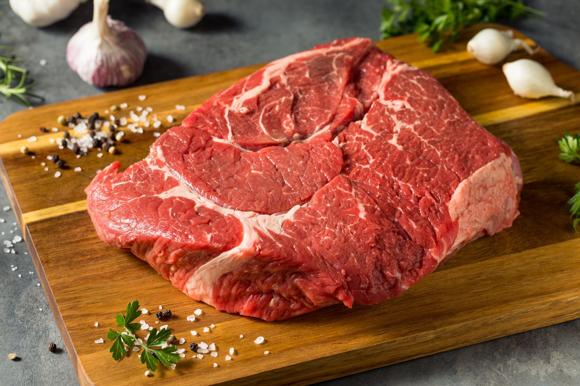 Raw chuck roast on a wooden cutting board with garlic, herbs, salt, and pepper.
