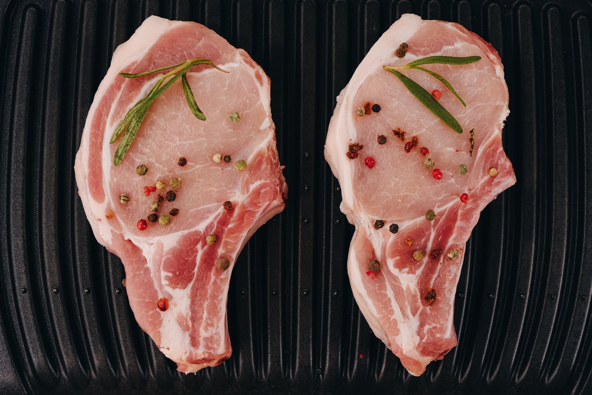 Two seasoned pork chops on a black grill, garnished with rosemary sprigs and peppercorns.