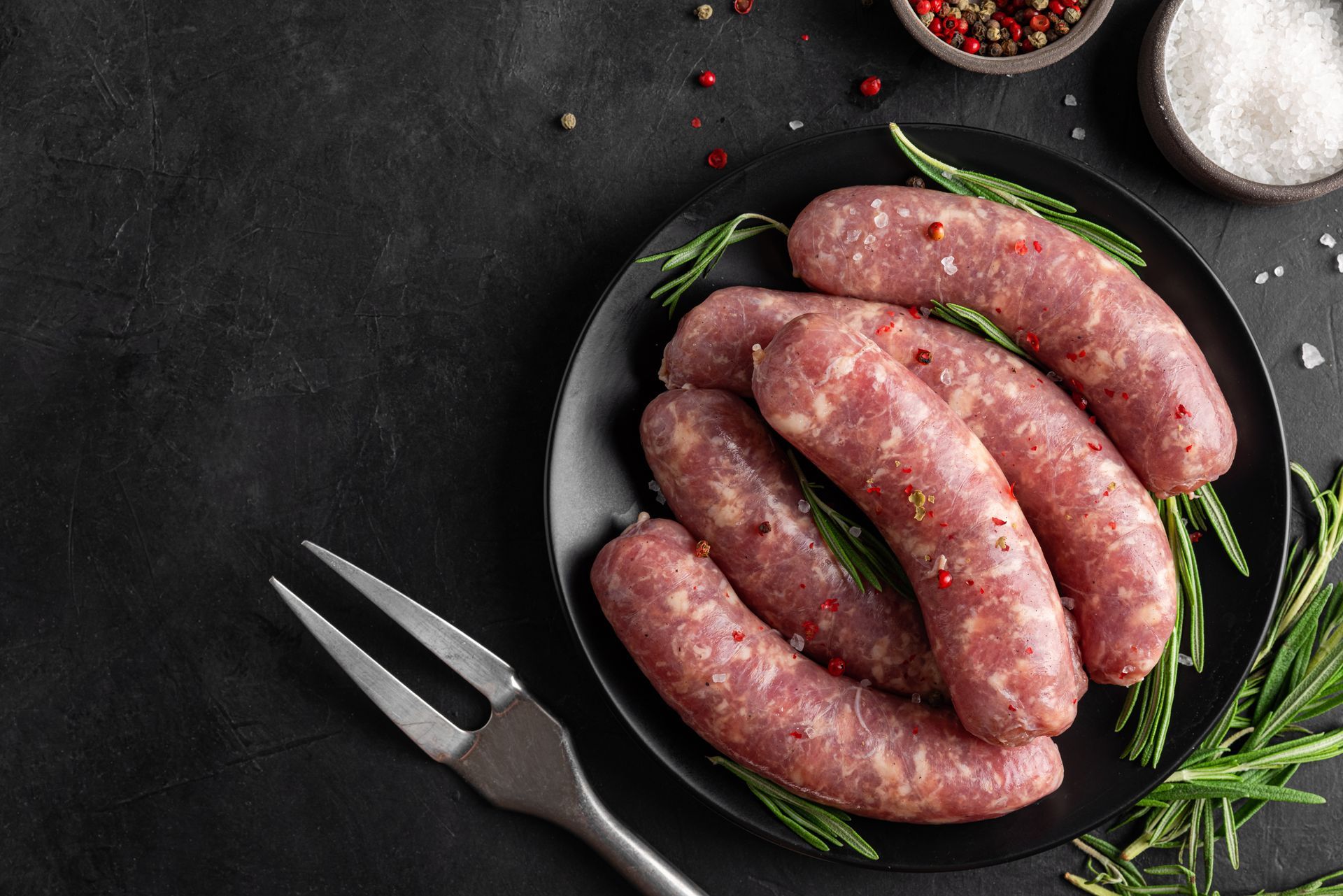 Raw sausages on a black plate with rosemary, a fork, and spices.