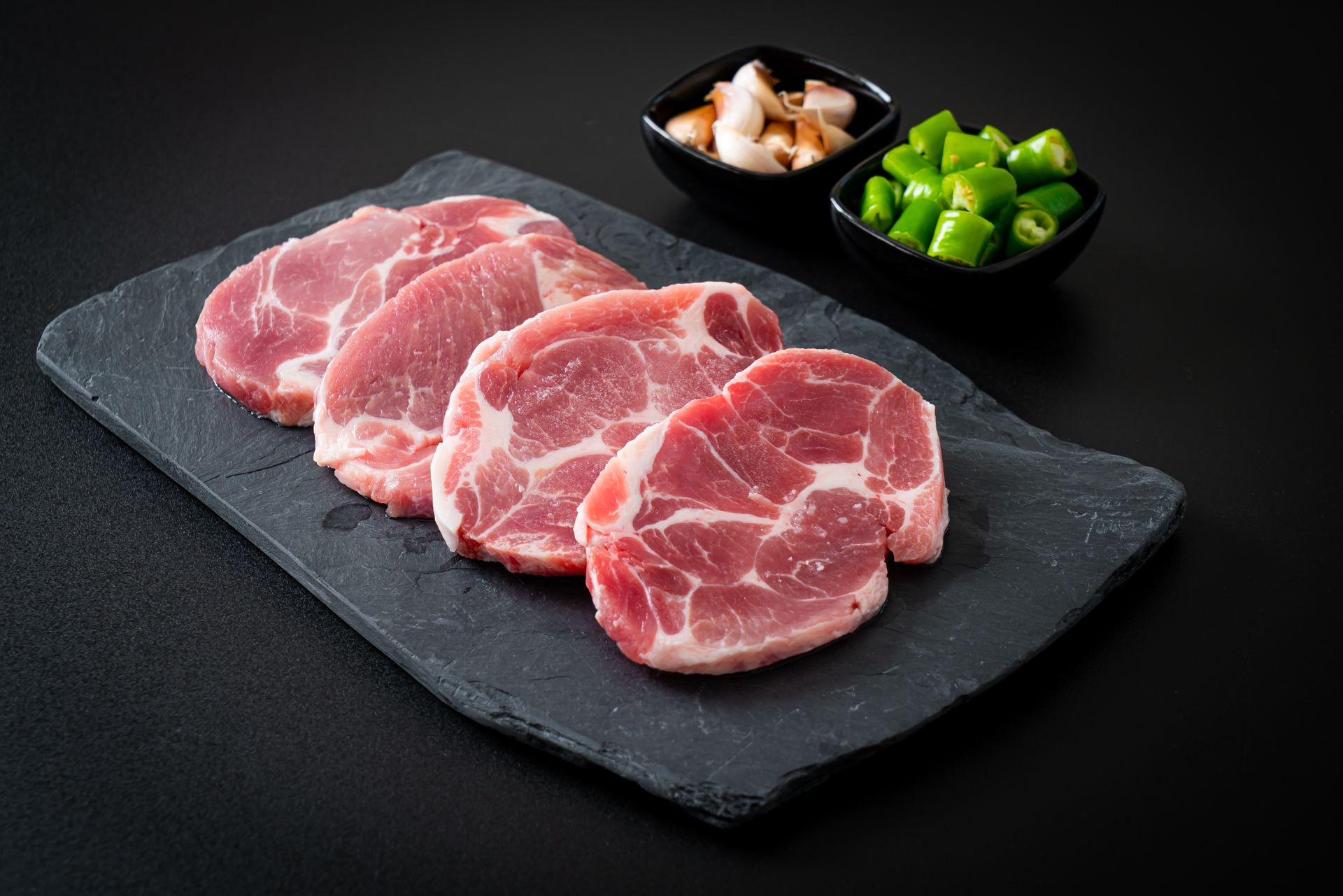 Slices of raw pork on a dark gray slate, with small bowls of green peppers and pickled vegetables.