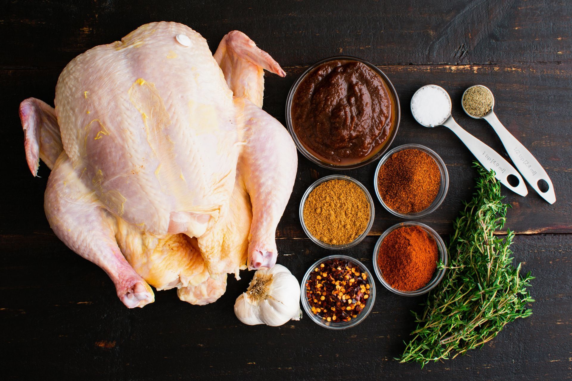 Raw whole chicken with various spices and sauces on a dark wooden surface.