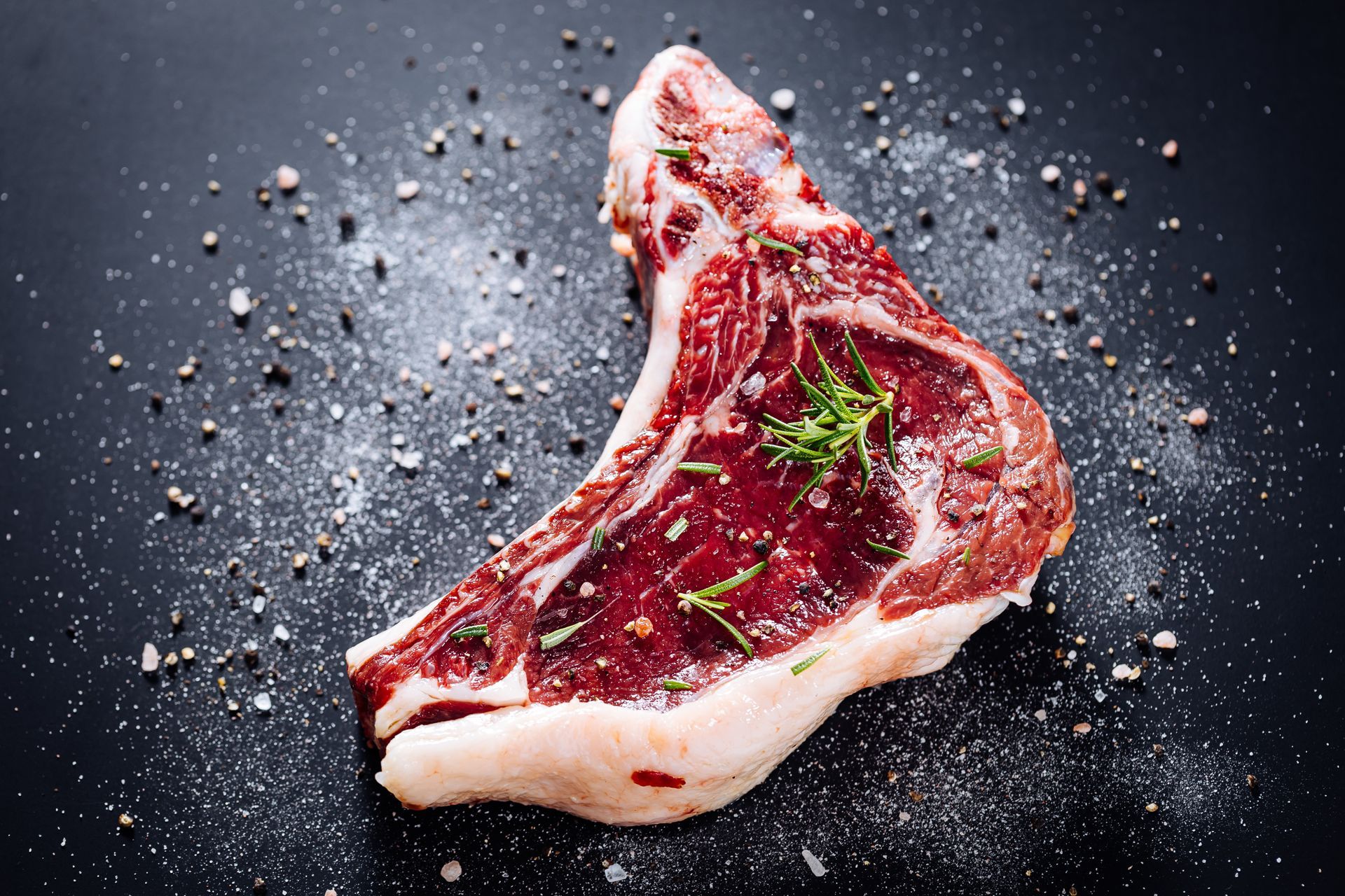 Raw, seasoned T-bone steak on a black surface sprinkled with salt and pepper.