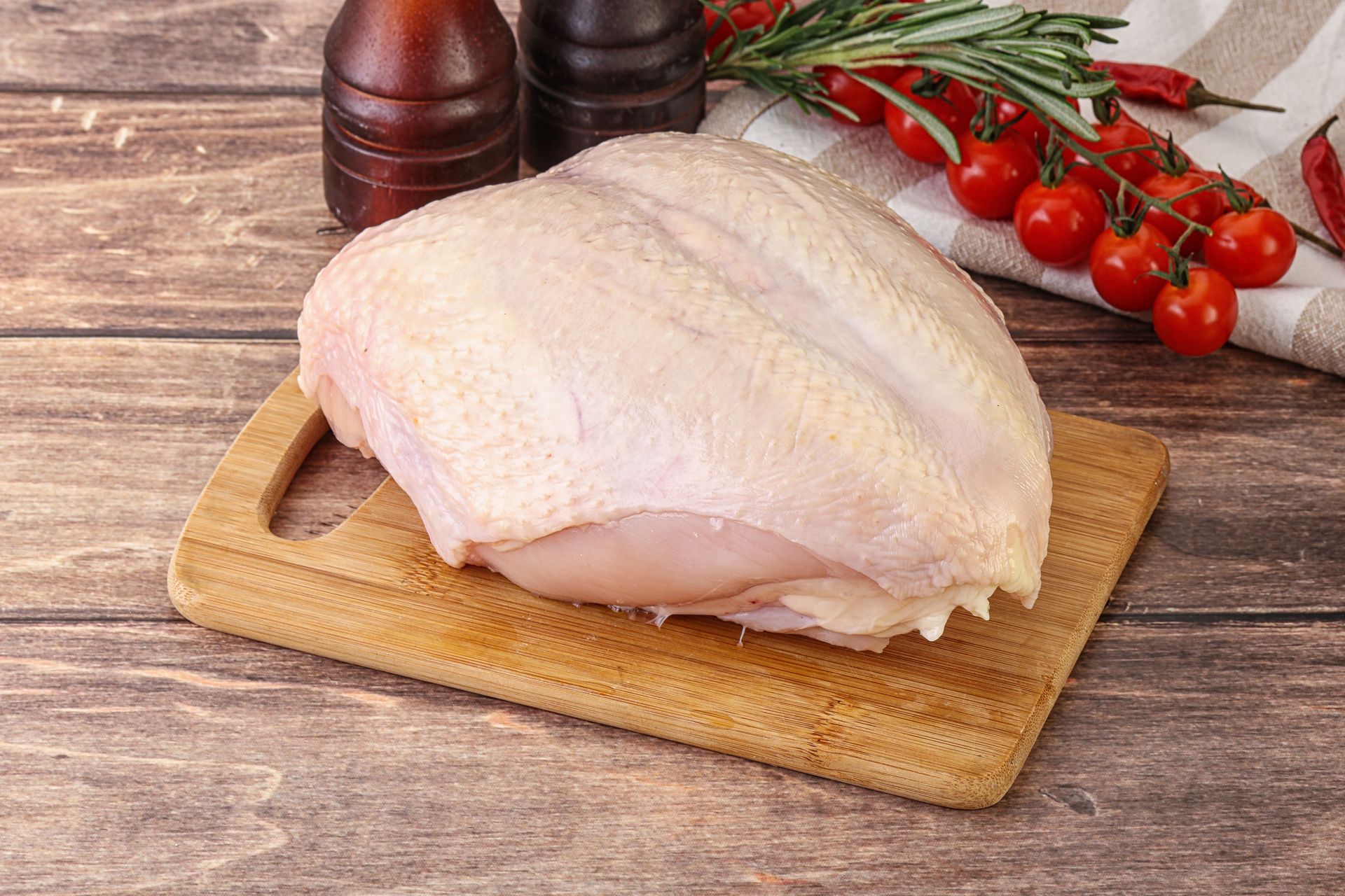 Raw chicken breast on a wooden cutting board, with tomatoes, rosemary, and peppercorns in the background.