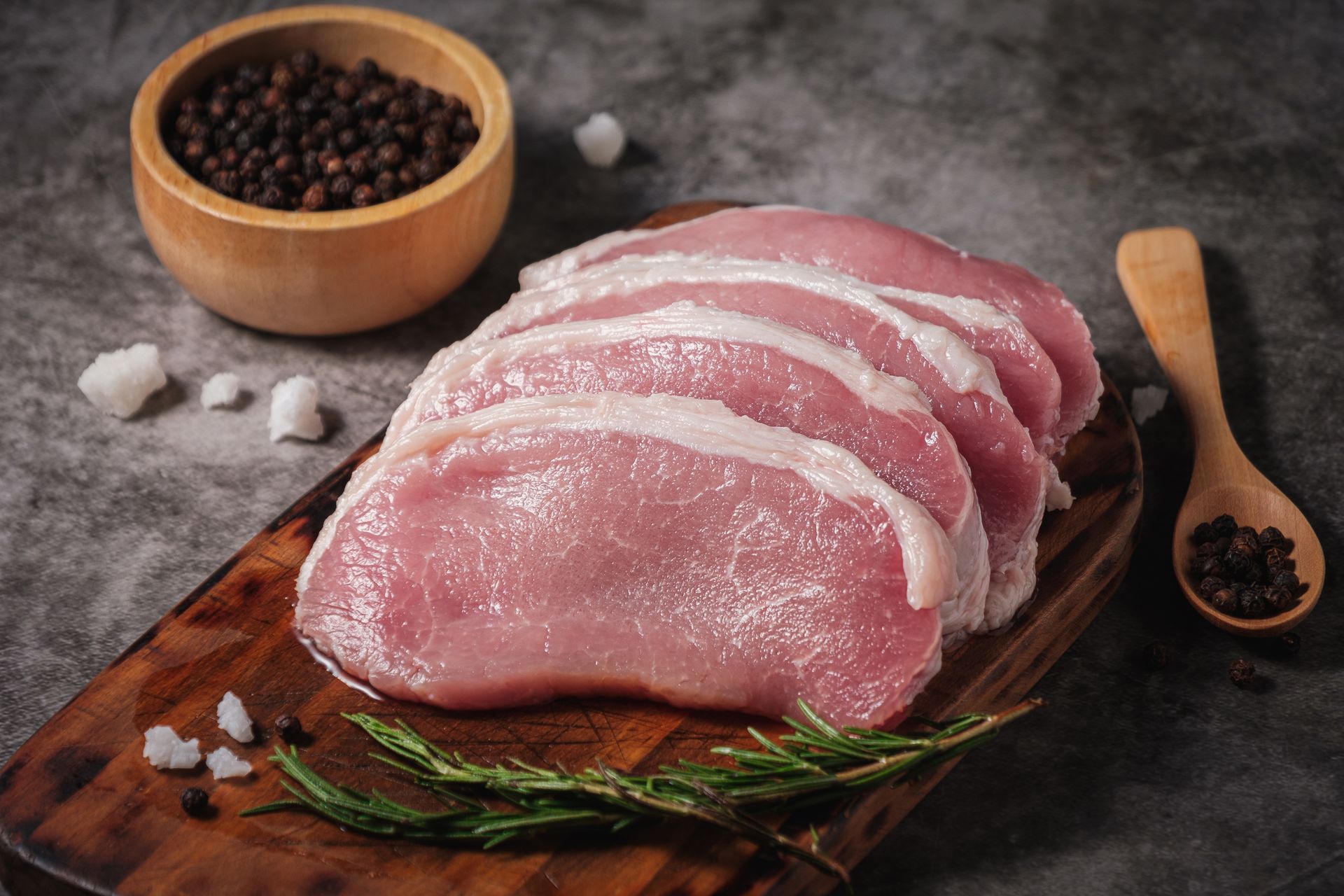 Pork chops on a wooden board with peppercorns, salt, and rosemary on a gray background.