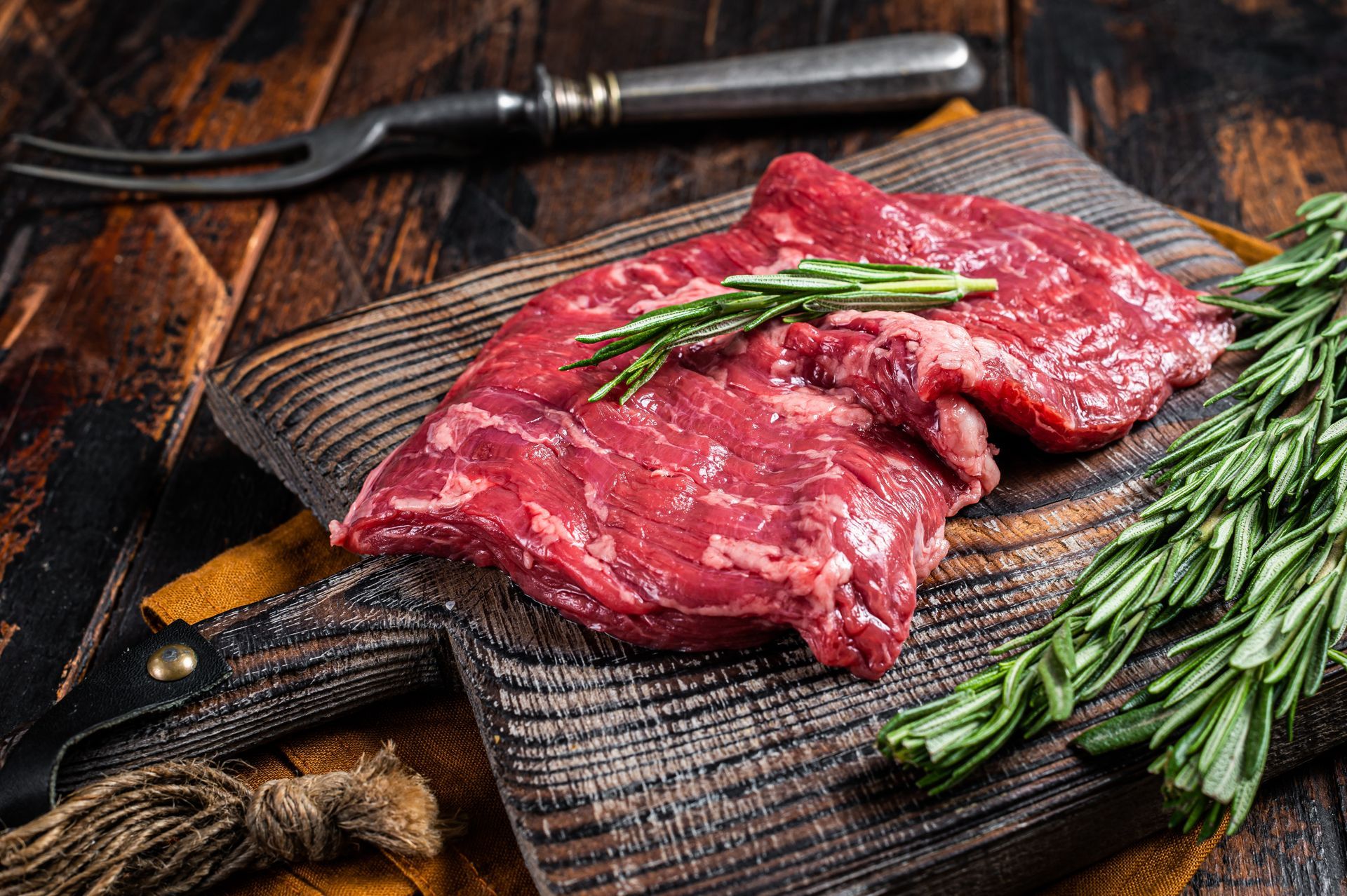 Raw flank steak on a wooden cutting board with rosemary sprigs and a meat fork.