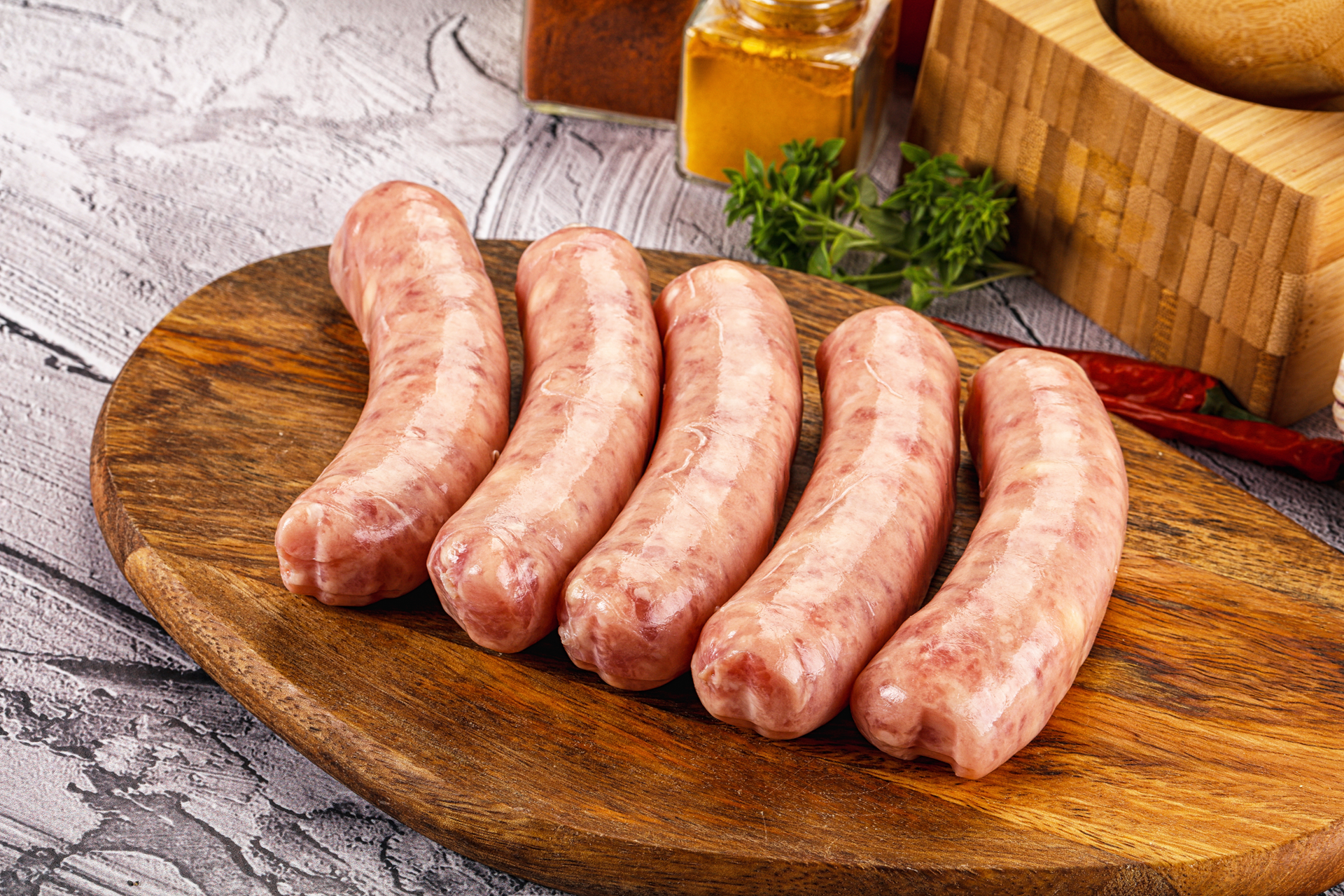 Five raw sausages on a wooden cutting board, with spices and herbs in the background.