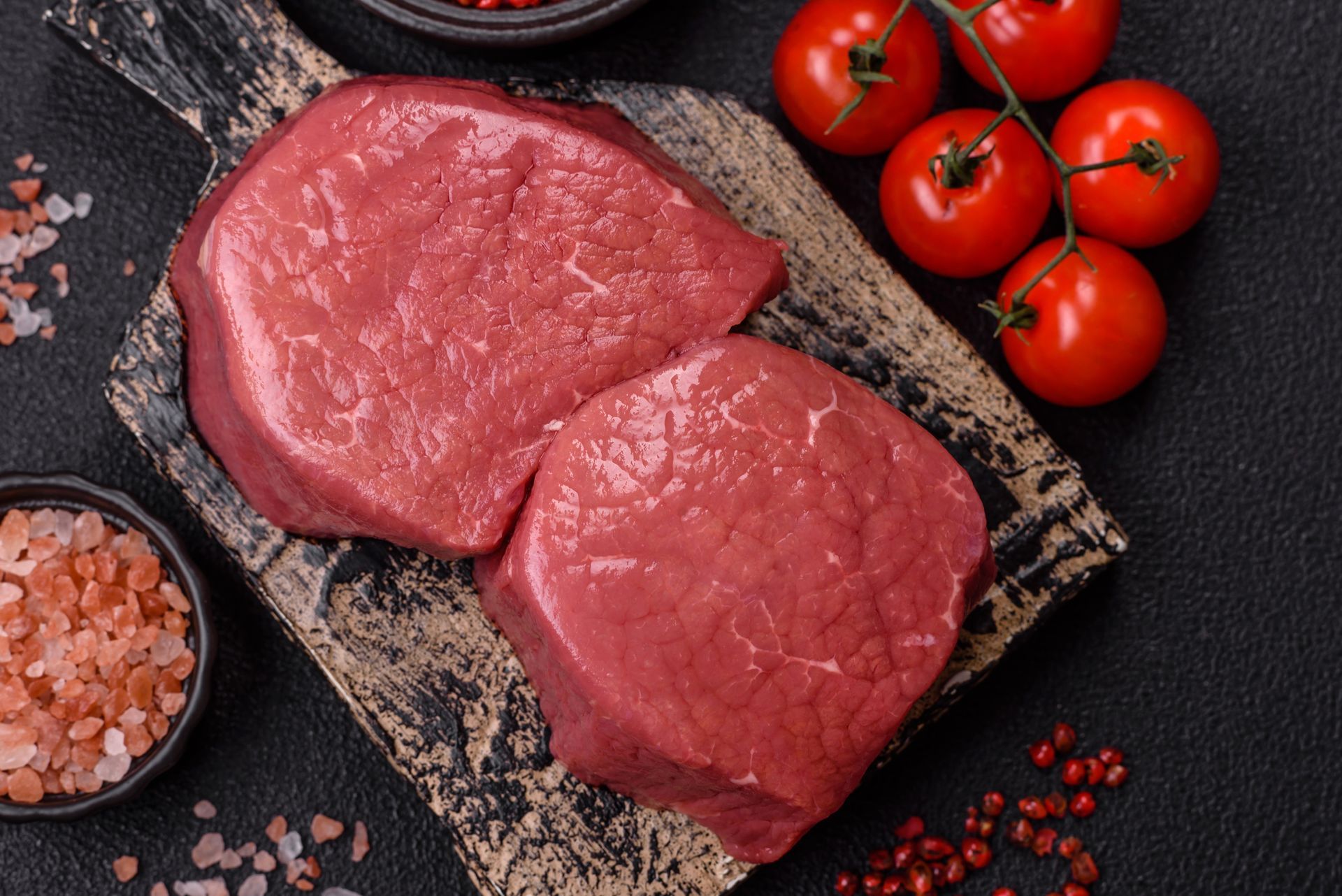 Two raw beef tenderloin steaks on a wooden cutting board with tomatoes and salt.