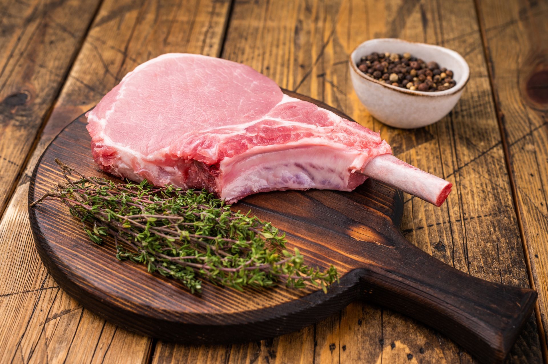 Pork chop on a wooden cutting board with thyme and peppercorns.